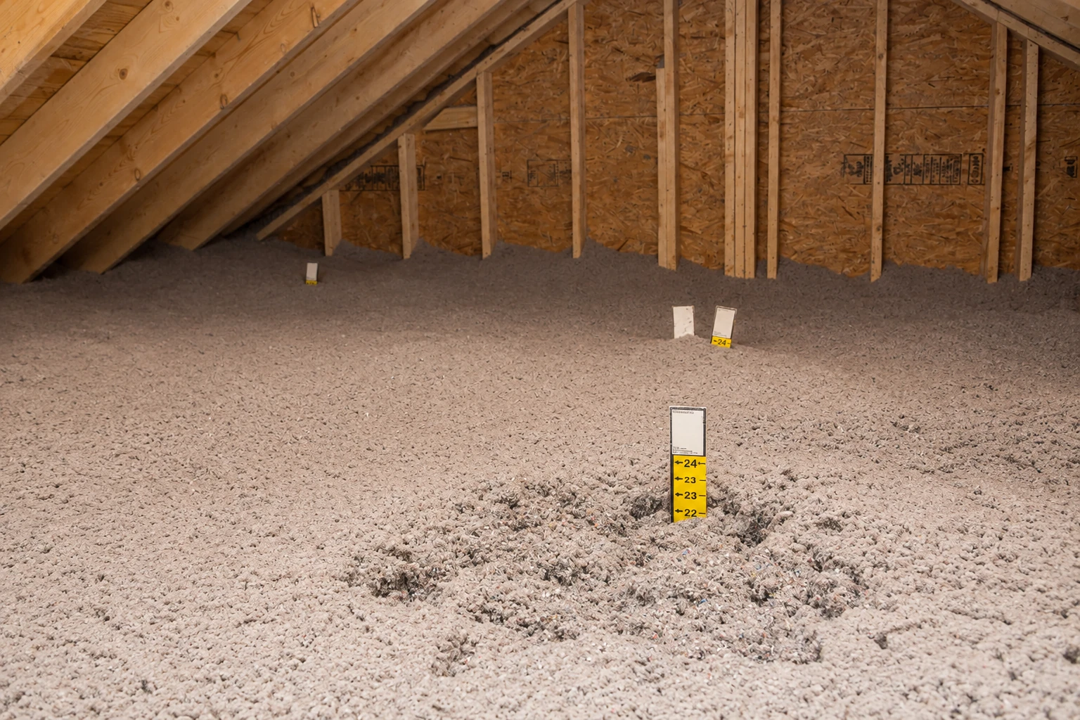 Blown-in insulation covers an attic floor beneath exposed wooden rafters, with depth markers protruding from the fill.