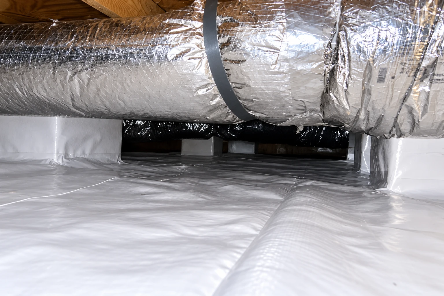 A low crawl space has a reflective metal duct overhead and a white vapor barrier covering the floor and walls.