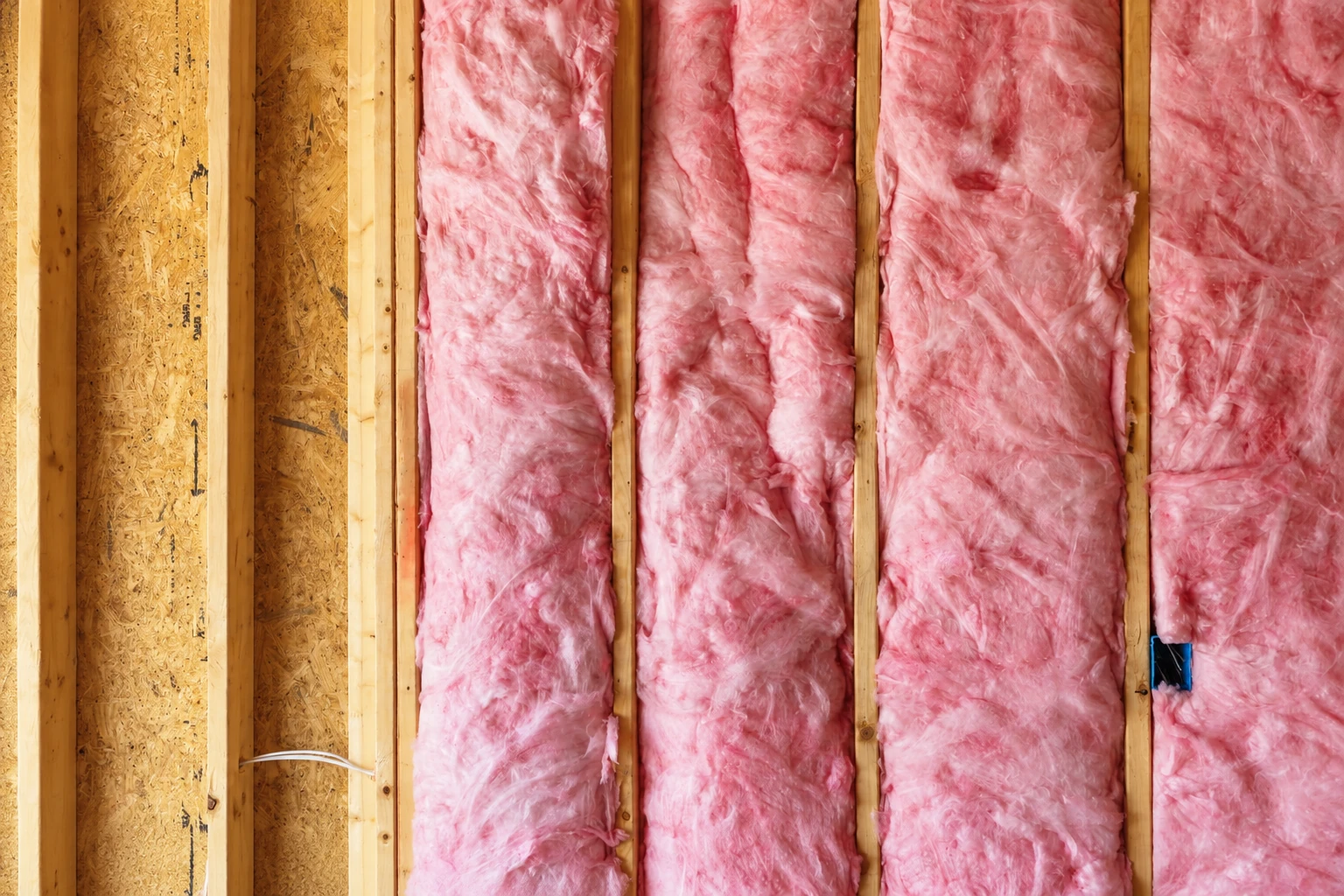 Pink batt insulation fills the spaces between wooden studs in an unfinished wall.