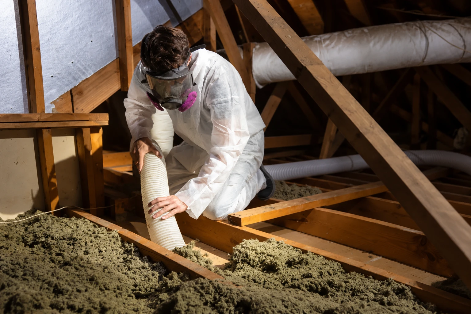 A worker in protective coveralls and a respirator uses a large hose to remove loose attic insulation.