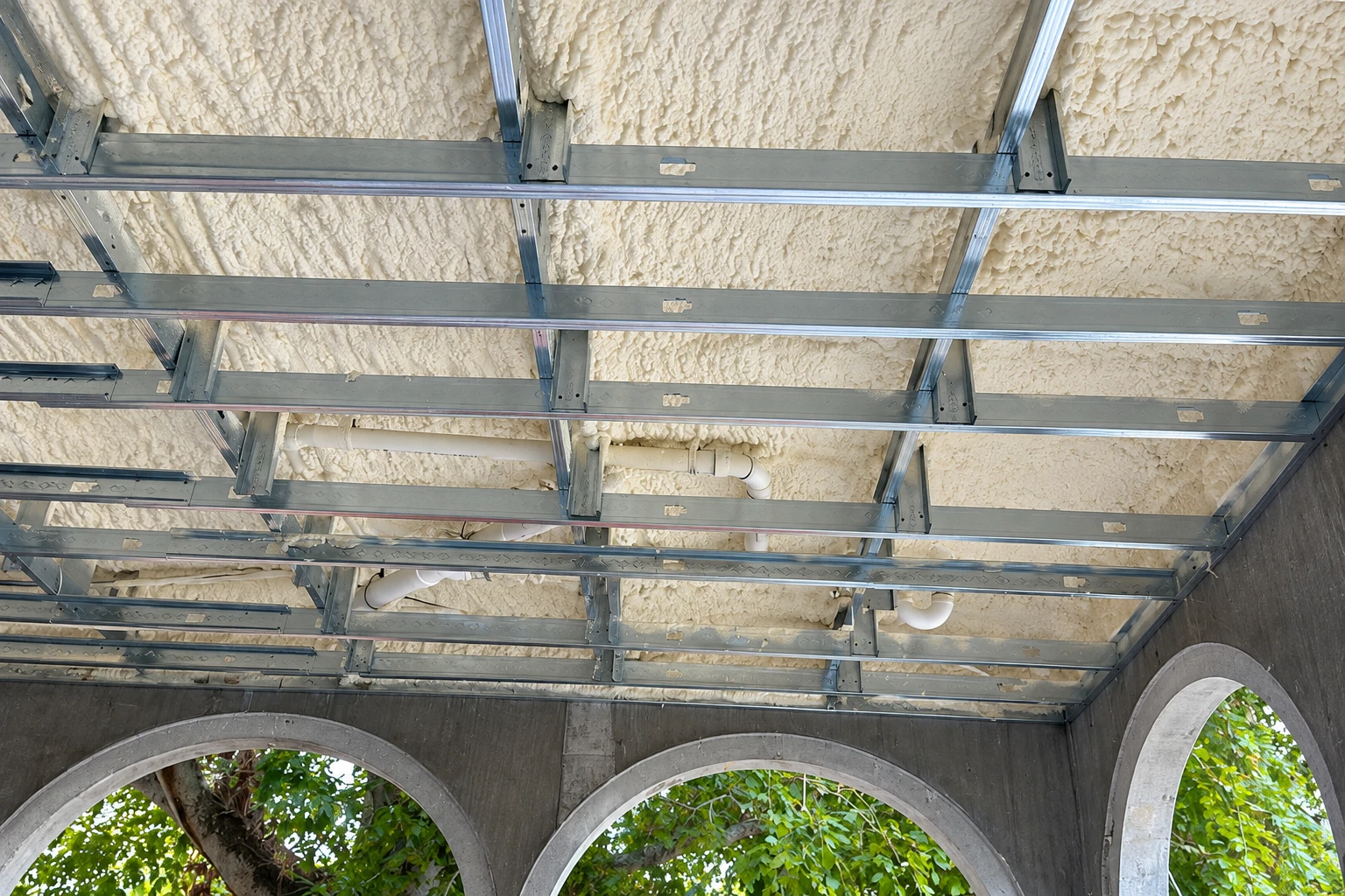 Spray foam insulation covers a ceiling behind a grid of metal framing, with arched openings and greenery below.