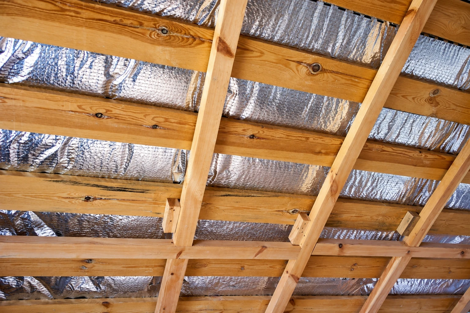Reflective foil vapor barrier is installed between wooden rafters on a sloped roof.