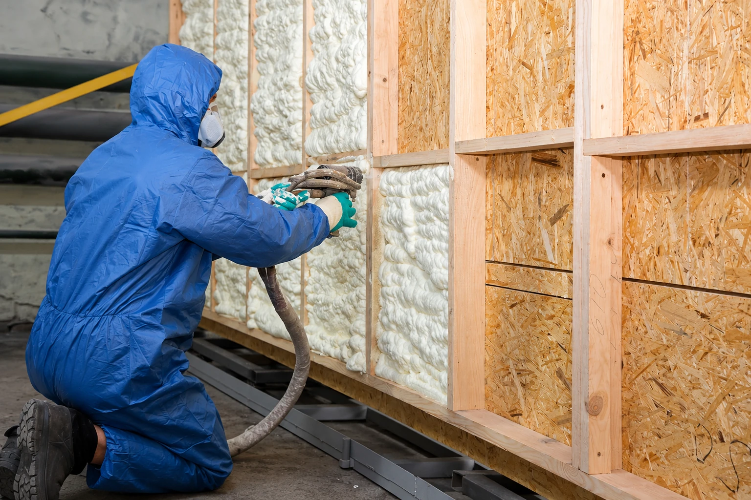 A worker in blue protective gear sprays foam insulation into the open bays of a framed wall.